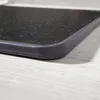 A close-up view of the corner of a black cutting board with a granite-like texture, resting on a light-colored wooden surface. Náhľad obrázku produktu