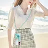 A woman wearing a white blouse and a plaid skirt is standing on a beach, with a phone case with a lanyard around her neck. Náhled obrázku produktu