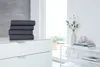 A stack of folded dark gray sheets on a white dresser, with a window and a shelf with decorative items in the background. Produktbild-Vorschau