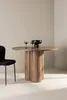 A round wooden dining table with a fluted pedestal base, set with two decorative glasses and a black bowl. A black chair is partially visible on the left. Produktbild-Vorschau