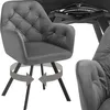 A gray upholstered armchair with a tufted back and arms, shown from the front and side. The armchair has a swivel base indicated by arrows. The base of a table is visible in the background. Produktbild-Vorschau
