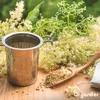 A metal tea infuser sits on a wooden surface surrounded by elderflower blossoms and a wooden spoon filled with dried elderflower. Náhľad obrázku produktu