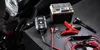 A motorcycle battery charger and accessories are laid out on a textured black surface, with a portion of a motorcycle visible in the background. Podgląd zdjęcia produktu