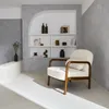 A cream-colored armchair with a bouclé fabric and a walnut-colored wooden frame is placed in a minimalist room with gray textured walls and a white floor. A built-in shelf unit with decorative items is visible behind the chair. Produktbild-Vorschau