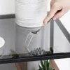 A hand holding a fork over a glass table with a vase and plants in the background. Produktbild-Vorschau