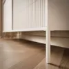 A close-up view of the bottom part of a beige cabinet with vertical slats and legs, resting on a wooden floor. Produktbild-Vorschau