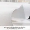 A close-up view of white fabric, likely a mattress protector or bedding, with text overlay describing its features. Produktbild-Vorschau