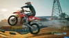 A motocross rider in mid-air on a red dirt bike, with a cityscape and industrial structures in the background. Produktbild-Vorschau