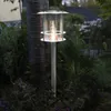 A solar-powered path light is illuminated in a garden at dusk, surrounded by plants and a tree trunk. Produktbild-Vorschau