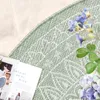 A close-up of a round outdoor rug with a geometric pattern, placed on a tiled floor. A magazine is open on the rug, and flowers are arranged on top of the rug. Produktbild-Vorschau