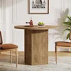 A round wooden dining table with a fluted base, set with two glasses of red wine, a bottle of wine, and a bowl of green apples. Two chairs with brown leather seats and cane backs are positioned on either side of the table. A framed picture hangs on the wal Produktbild-Vorschau