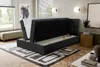 A bedroom scene with a black boxspring bed with its storage compartment open, revealing white bedding. A rug with a geometric pattern is on the floor, and a bench is visible in the background. Voir les images du produit