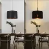 A split image showing a black pendant lamp with a textured fabric shade and a gold interior, displayed in a dining room setting with a wooden table and chairs. The left side shows the lamp unlit against a white wall, while the right side shows it lit, cast Anteprima dell’immagine prodotto