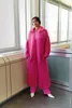 A woman stands in front of a window, wearing a long pink coat and pink pants. Produktbild-Vorschau