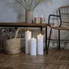 Three LED candles are lit on a wooden floor in front of a rustic bench with a basket of bottles and a chair in the background. Produktbild-Vorschau