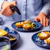 A person is using a culinary torch to caramelize the tops of small tarts on a blue plate. Voir les images du produit