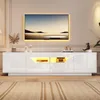 A modern white TV stand with LED lighting below the shelves is shown in a room with a large television mounted on the wall above it. The TV stand is on a carpeted floor. Produktbild-Vorschau