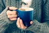 A person wearing a gray knitted sweater holds a blue mug with steam rising from it. Produktbild-Vorschau