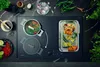 A black induction cooktop with two pots and a grilling tray with food. Ingredients like garlic, chili peppers, and herbs are arranged around the cooktop. Voir les images du produit