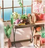 A close-up view of a miniature dollhouse scene featuring plants, a table, and decorative items. Náhled obrázku produktu