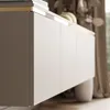 A close-up view of a modern TV cabinet with a sleek design, featuring multiple doors and a subtle gold trim along the top edge. The cabinet is shown in a well-lit interior setting with soft curtains in the background. Produktbild-Vorschau