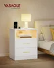 A white nightstand with two drawers and an open shelf, placed next to a bed. A lamp, phone, and books are on the nightstand. Náhled obrázku produktu