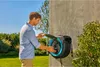 A man is interacting with a wall-mounted hose reel in a garden setting. Voir les images du produit