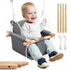 A baby sitting in a gray fabric swing with wooden bars, suspended by white ropes. The baby is smiling and wearing a light blue long-sleeved shirt and light blue pants with dark blue sneakers. Several wooden dowels and two sets of rope with metal rings are  Produktbild-Vorschau