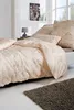 A neatly made bed with beige and gray patterned bedding, a bedside table with a lamp, and a framed picture on the wall. Produktbild-Vorschau