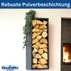 A black metal firewood rack filled with logs, mounted on a wall next to a fireplace. A green vine hangs from the top of the rack. Produktbild-Vorschau