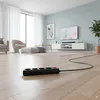 A power strip with multiple outlets and a cable is placed on a wooden floor in a living room setting. The room has a sofa, armchair, coffee table, and a TV on a wall-mounted unit. Náhled obrázku produktu