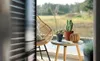 A balcony scene with a chair, table, and plants, viewed through blinds. Produktbild-Vorschau
