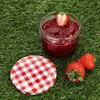 A jar of strawberry jam with a red and white checkered lid and strawberries on grass. Produktbild-Vorschau