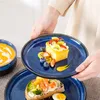 A person is holding a plate with a slice of cake topped with fruit, and another plate with an open-faced sandwich is visible below. Voir les images du produit