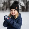 A young girl wearing winter clothing, including a navy blue jacket, black hat with a pom-pom, and navy blue mittens with snowman designs, stands in a snowy environment with falling snow. Náhled obrázku produktu