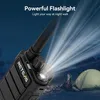 A walkie-talkie is shown in a dark outdoor setting with a tent and mountains in the background. The walkie-talkie's flashlight is on, illuminating the scene. Anteprima dell’immagine prodotto
