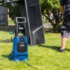 A person is using a blue and black pressure washer to clean a large, dark outdoor furniture piece in a grassy yard. Produktbild-Vorschau
