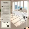 A white massage table set up in a bright, modern room with large windows overlooking a scenic view. Náhled obrázku produktu
