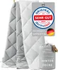 Two quilted blankets are displayed, one gray and one white, with a marketing tag and a packaged blanket in the foreground. Produktbild-Vorschau