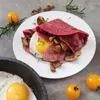 A breakfast dish with a pink crepe, fried egg, bacon, and mushrooms, served on a white plate, with cherry tomatoes and rosemary sprigs scattered around. Produktbild-Vorschau