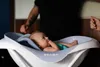 A baby is lying in a bathtub with a person's hands visible. The baby is looking up and has a finger in their mouth. Anteprima dell’immagine prodotto