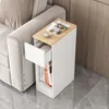 A narrow white bedside table with a wooden top is placed next to a sofa. The table has an open drawer and a shelf filled with books. A cup and magazines are on top of the table. Produktbild-Vorschau