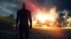 A bald man in a dark suit stands in front of a burning vehicle on a sandy beach. Náhľad obrázku produktu