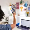 A person is holding up a decorative letter 'S' in a brightly lit room with various decorative items on the wall and shelves. Produktbild-Vorschau