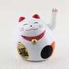 A white Maneki-neko (waving cat) figurine with red ears and collar, holding a gold coin with Chinese characters, sits on a patterned surface. Produktbild-Vorschau