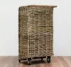 A wicker laundry basket with wheels on a wooden floor against a white wall. Produktbild-Vorschau