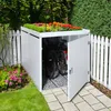 A white metal bicycle garage with a green roof garden, containing two bicycles, is situated in a garden next to a white picket fence. Náhled obrázku produktu