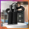 A black jump rope with foam handles is displayed on a white pedestal against a blurred background. Podgląd zdjęcia produktu