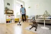 A man is vacuuming a living room while his dog lies on a rug nearby. The room has a couch, a coffee table, a bookshelf, and a vintage radio. Anteprima dell’immagine prodotto