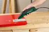 A person's hand is holding an electric scraper and using it on a red metal surface. Produktbild-Vorschau